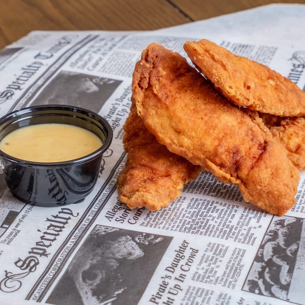 Crispy golden fried chicken tenders arranged beside a cup of creamy dipping sauce on newspaper-lined paper.