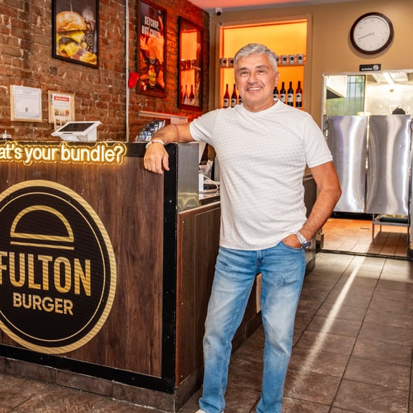 Smiling man stands beside the Fulton Burger restaurant counter, featuring a glowing “what’s your bundle?” sign and exposed brick walls.