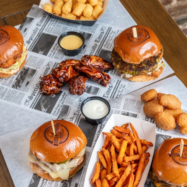 Assorted burgers, barbecue chicken wings, tater tots, dipping sauces, and sweet potato fries arranged on newspaper-lined trays.