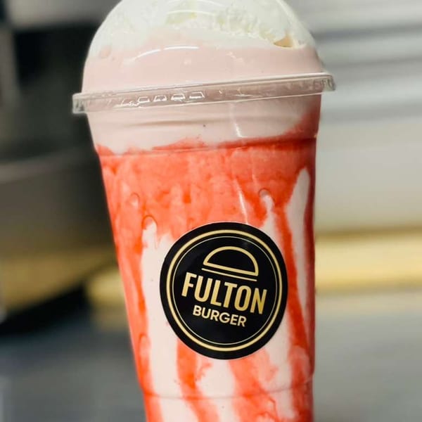 Strawberry milkshake from Fulton Burger, topped with whipped cream, in a clear plastic cup.