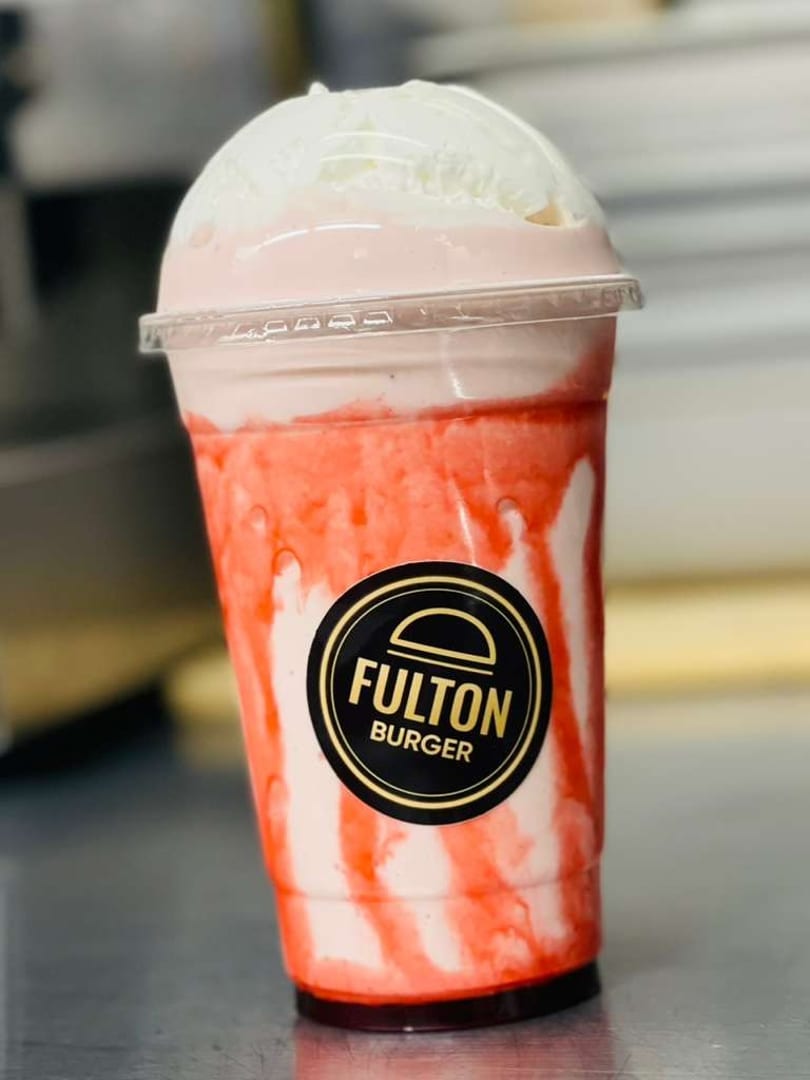 Strawberry milkshake from Fulton Burger, topped with whipped cream, in a clear plastic cup.