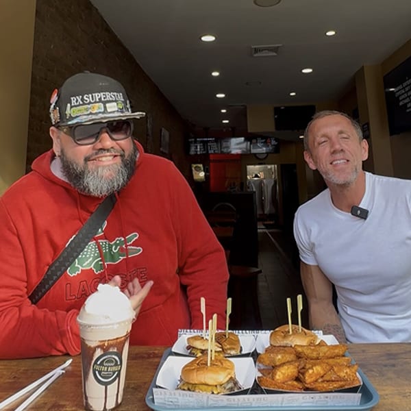Two men smiling with burgers, fries, and a milkshake on a table in a restaurant.