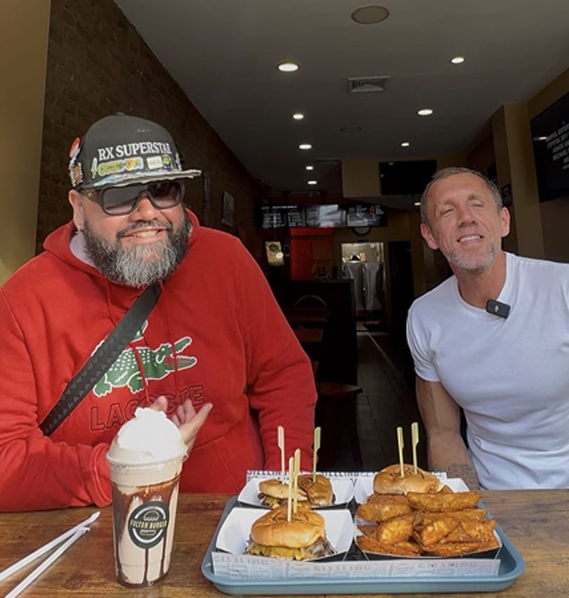 Two men smiling with burgers, fries, and a milkshake on a table in a restaurant.