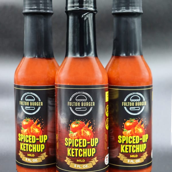 Three bottles of Fulton Burger Spiced-Up Ketchup, mild, 5 fl. oz., on a dark background.