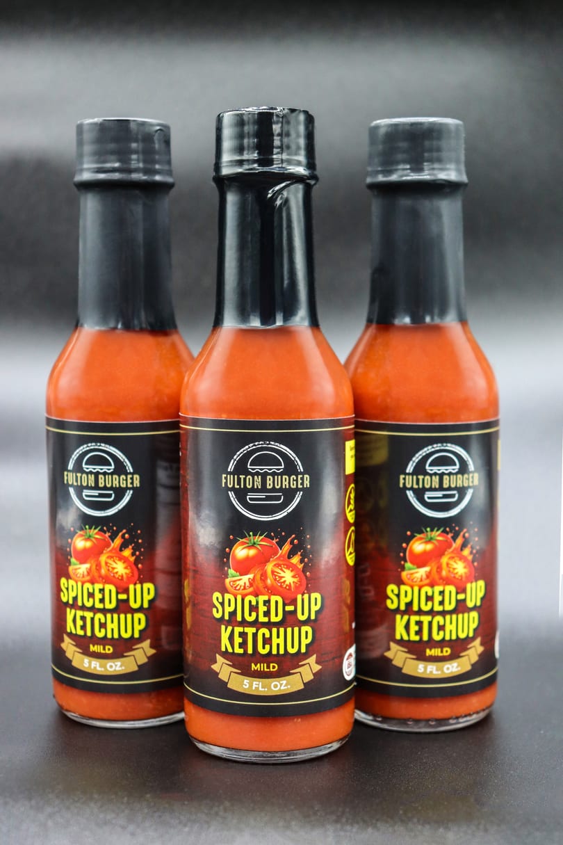 Three bottles of Fulton Burger Spiced-Up Ketchup, mild, 5 fl. oz., on a dark background.