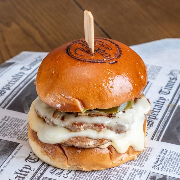 Double cheeseburger with melted cheese, pickles, and grilled onions on a glossy bun, secured with a wooden skewer.