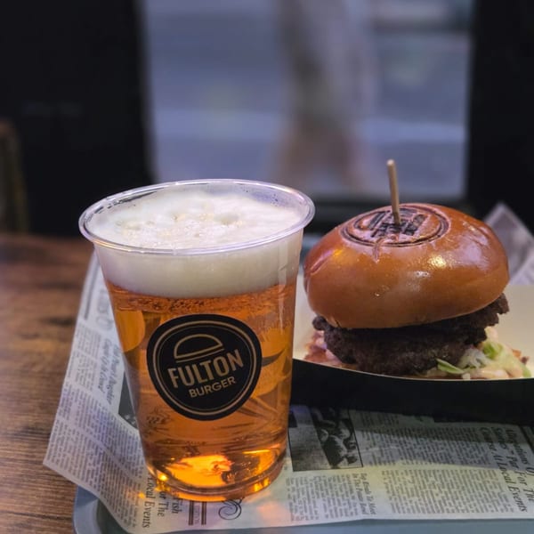 Fulton Burger and a beer in a branded plastic cup, served on a tray with newspaper lining.