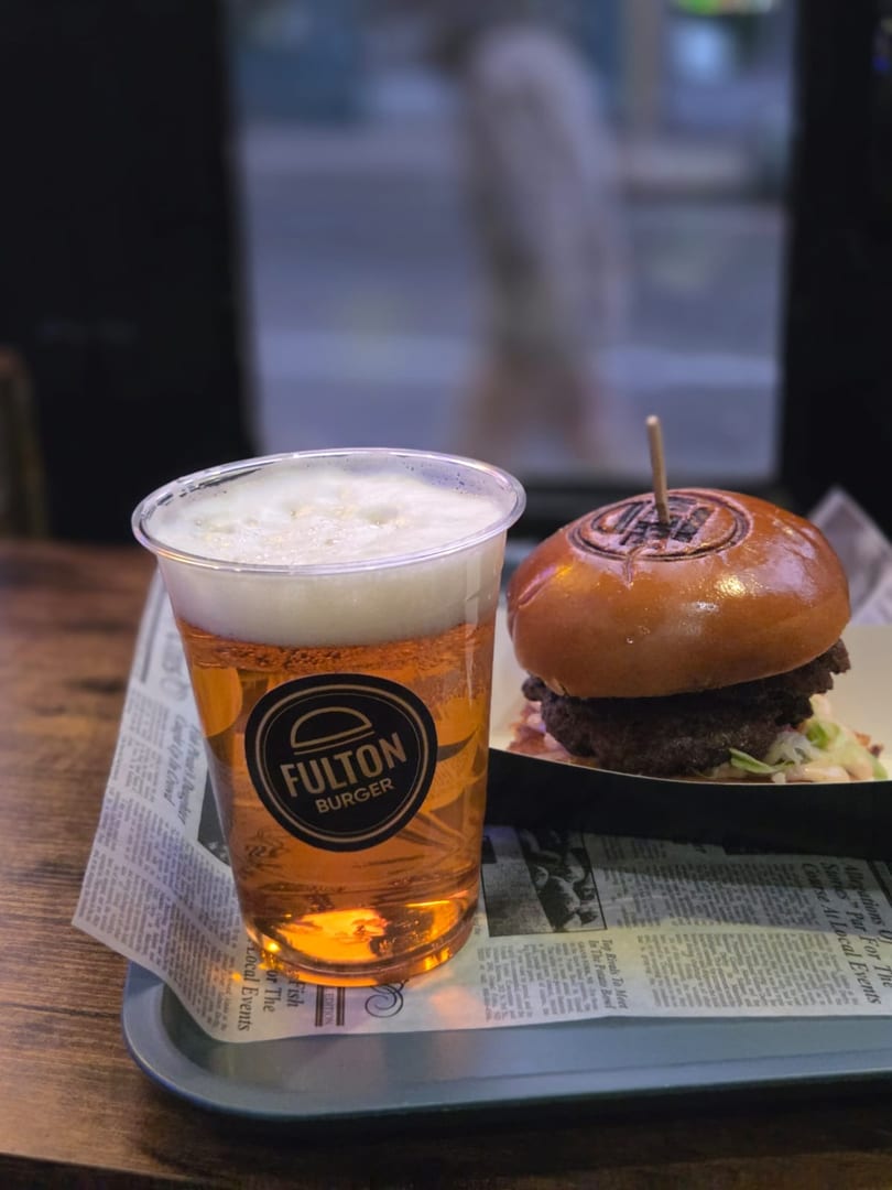 Fulton Burger and a beer in a branded plastic cup, served on a tray with newspaper lining.