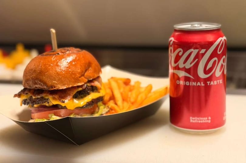 A double cheeseburger with bacon, lettuce, tomato, and fries, next to a Coca-Cola can.