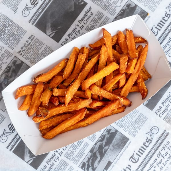 Crispy golden sweet potato fries piled in a white paper tray on newspaper-style restaurant wrapping.