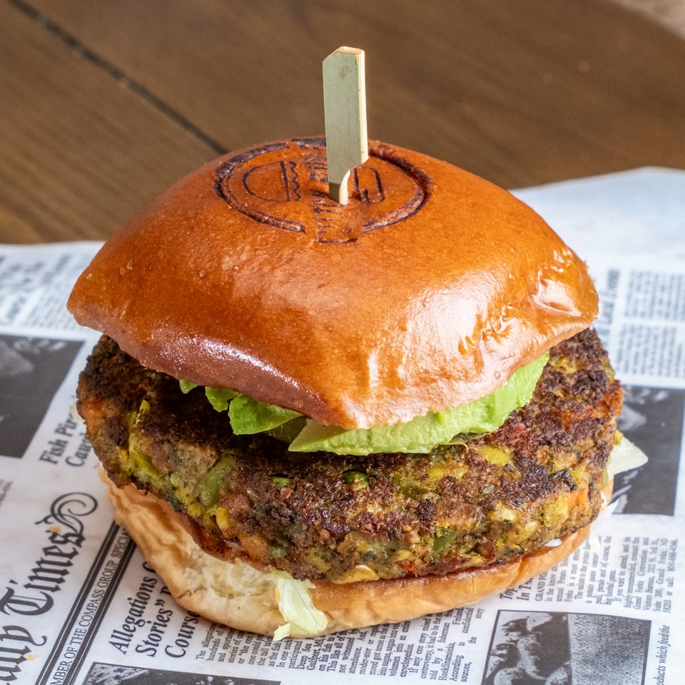 Close-up of a veggie burger with a glossy bun, lettuce, avocado, and a textured vegetable patty on newspaper-style paper.