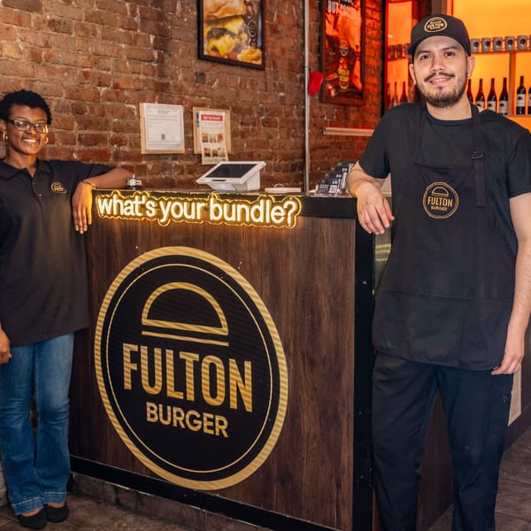 Two Fulton Burger staff members pose beside the restaurant counter, illuminated by a neon “what’s your bundle?” sign and orange-lit wine shelves.